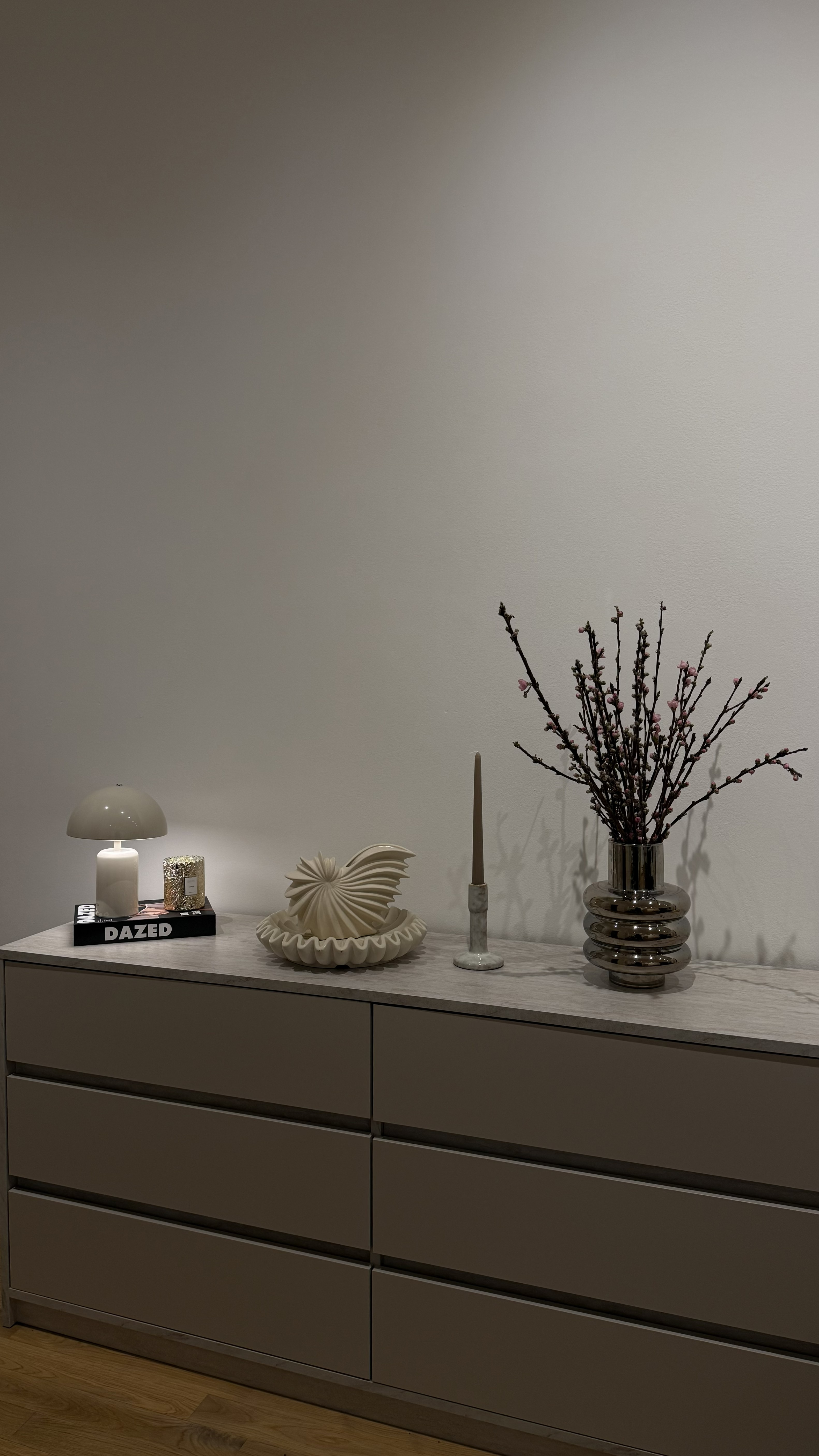 Eine elegante Kommode mit Kerzen, einem weißen Dekoobjekt und einem Ast in einer silbernen Vase