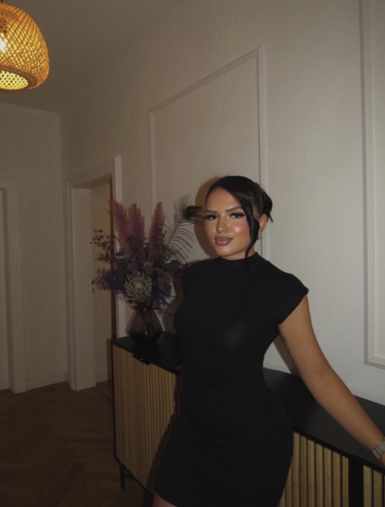Junge Frau mit elegantem schwarzen Kleid und Lächeln steht vor einer Vase mit Trockenblumen